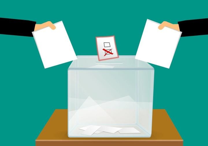 Symbolbild für Wahlen. Zwei Personen heben ihren Stimmzettel neben der Wahlurne.