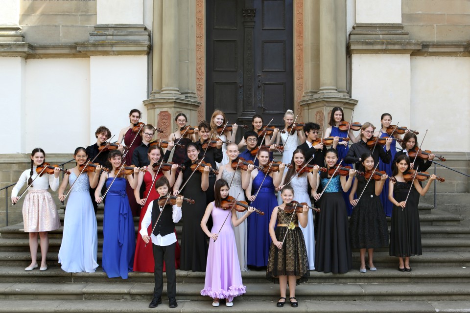 die Teilnehmerinnen und Teilnehmer am Violinenwettbewerb stehen mit ihren Violinen auf einer Treppe vor einer Eingangstüre zu einer Kirche. Alle sind schick für den Konzertabend gekleidet.