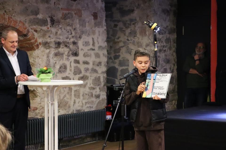 Junge steht an einem Mikrofon und zeigt ein Bild zu dem er etwas erklärt. Auf der linken Seite steht ein Mann an einem Stehtisch.