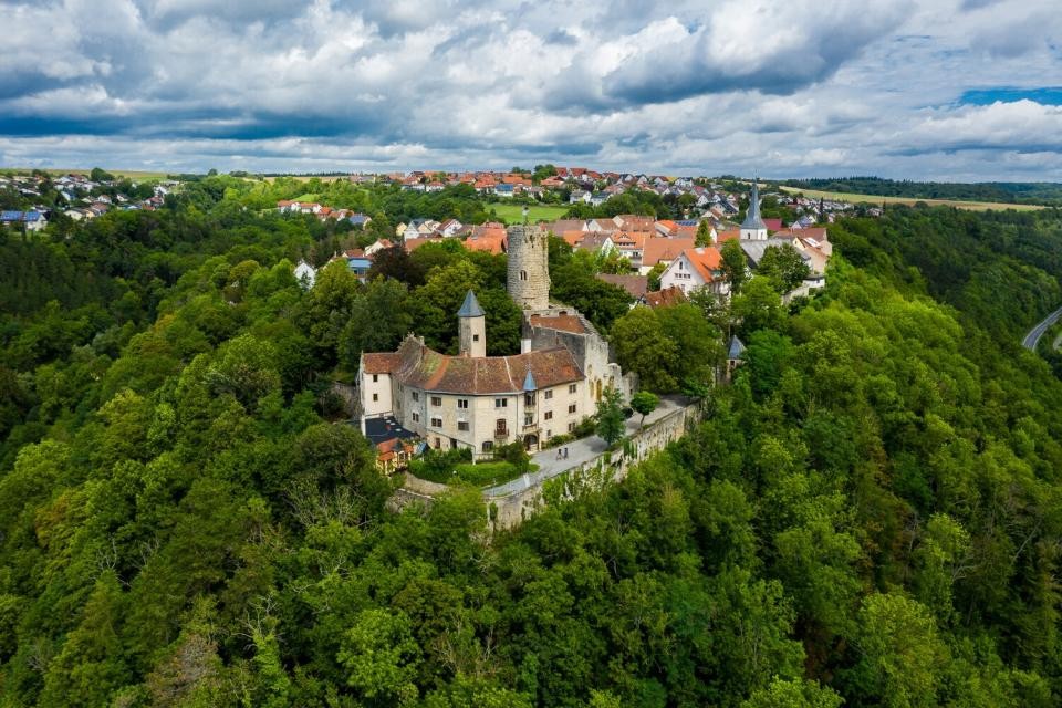 Eine Luftaufnahme der Burg Krautheim, die sich auf einer Anhöhe befindet. Drumherum sind  grün Bäme.Im Hintergrund befinden sich Häuser des Ortes und der Kirchturm ist zu sehen.