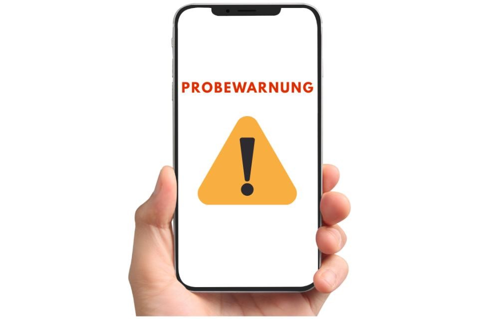 Hand, die ein Smartphone hält. Auf dem Smartphone ist in roter Schrift das Wort "Probewarnung", darunter ein gelbes Warnschild mit schwarzem Ausrufezeichen.