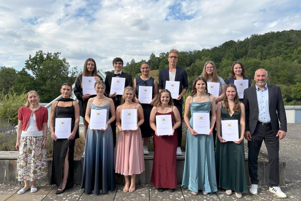 Schüler mit Zeugnissen in der Hand, rechts und links Lehrer im Freien, im Hintergrund Bäume