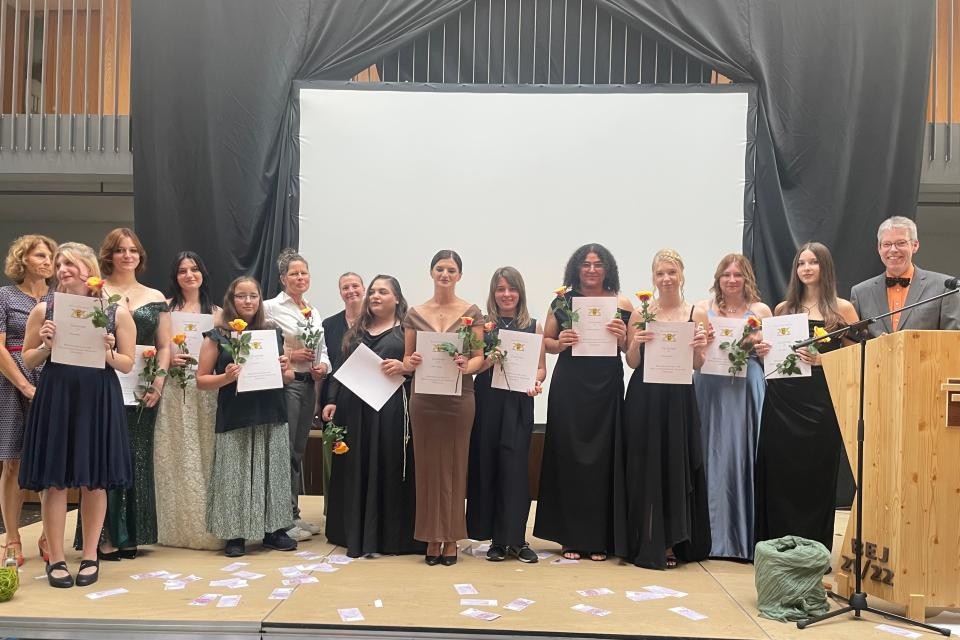 Absolventinnen auf einer Bühne mit Zeugnissen und Blumen in der Hand, links und rechts Lehrer, auf der rechten Seite befindet sich ein Holzpodest, im Hintergrund graue Vorhänge