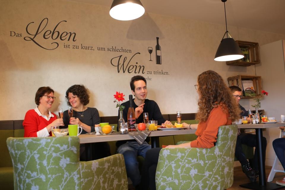 Eine gemütliche, moderne Gaststube in der 4 Personen gemeinsam frühstücken. An der Wand steht der Schriftzug "Das Leben ist zu kurz, um schlechten Wein zu trinken" von Goethe.