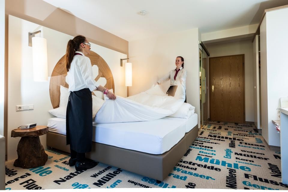Ein Doppelzimmer im Hotel. Zwei Mitarbeiterinnen in weißer Bluse und schwarzer Hose schütteln gemeinsam das Bett auf und richten das Zimmer.