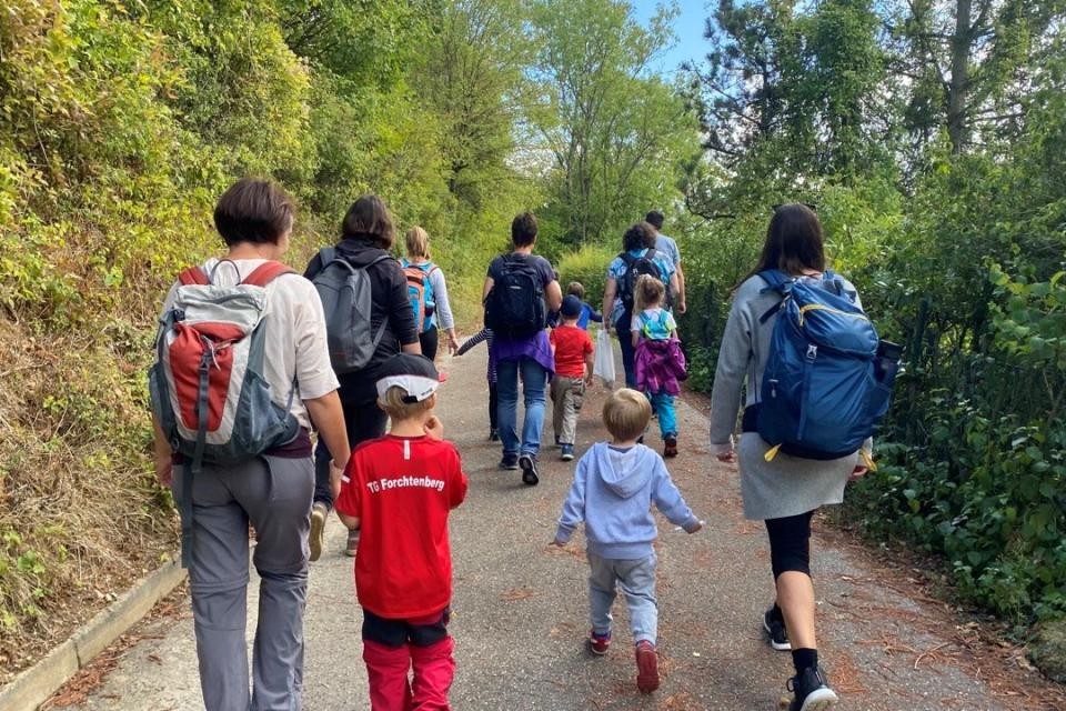 Kinder und Eltern laufen sind von hinten zu sehen, teilweise tragen sie Rucksäcke. Sie laufen auf einem asphaltierten Weg zwischen grünen Hecken.
