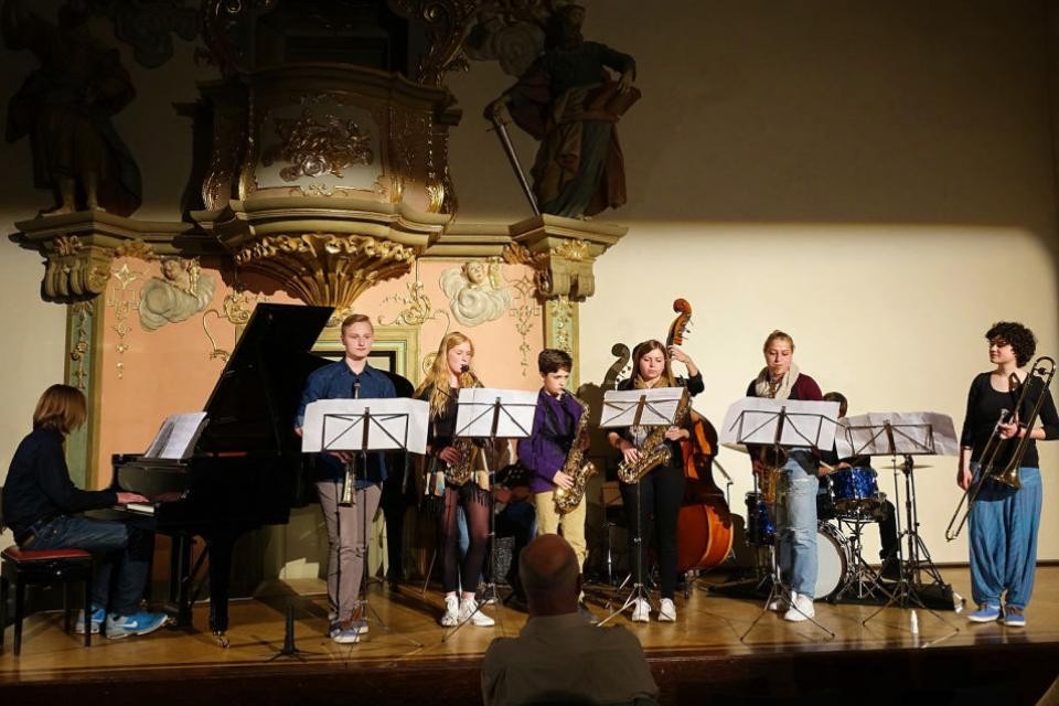 Links sitzt eine Frau am Flügel, daneben stehen 6 junge Musiker mit Klarinette und Saxophone. Auf der rechten Seite eine Person mit Posaune. Im Hintergrund spielt jemand sitzend Kontrabass. Vor den Personen stehen Notenständer mit weißen Notenblättern.
