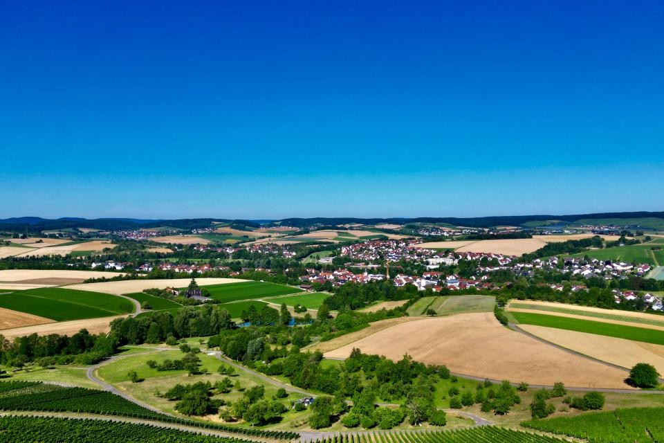 Gemeinde Bretzfeld Eine Drohnenaufnahme der Gemeinde Bretzfeld. Im unteren Teil sind Weinberge mit grünen Blättern zu sehen. Danach folgen Wiesen mit Bäumen und Sträuchern. In der Mitte sind Häuser der Gemeine Bretzfeld zu sehen. Drumherum Wiesen und Äcker, teilweise grün, teilweise braun/beige. Dazwischen immer wieder Baumstreifen. In der Ferne sind Hügel zu sehen. Der Himmel ist strahlend blau.