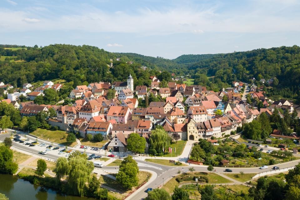Gemeinde Forchtenberg Vor bewaldeten Hügeln mit grünen Bäumen schmiegt sich das Städtchen Forchtenberg. In der Bildmitte ragt die Kirche mit dem Kirchturm hervor. Außenrum Fachwerkhäuser eingebettet in die Stadtmauer. Links unten fließt der Fluss Kocher, daneben parken einige Autos auf einem Parkplatz.