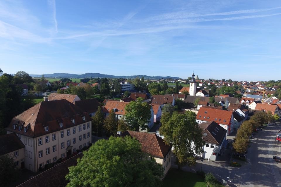 Gemeinde Kupferzell Blick von oben auf einen Teil der Stadt. Geprägt von grünen Bäumen, historischen Gebäuden, dem Kirchturm der über den Häuserdächern empor streckt. Im Hintergrund sieht man die bewaldeten Waldenburger Berge.