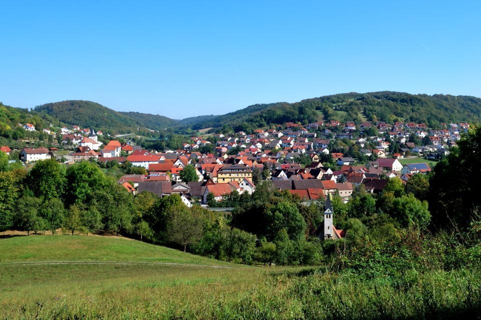 Einige Häuser zwischen grünen bewaldeten Hügeln stellt die Gemeinde Mulfingen dar. In der Mitte sticht ein gelbes älteres Bauwerk hervor. Im Vordergrund ist eine grüne Wiese und vor dem Ort ein kleiner Kapellenturm. Der Himmel ist hellblau.