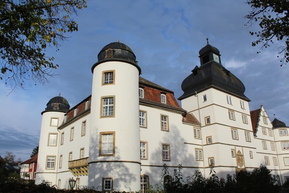 Das weiße Schloss aus der Renaissance mit einer zwei Seiten Ansicht. Zu sehen sind 2 runde und ein eckiger Turm. Die Dächer der Türme sind schwarz und abgerundet. das Dach des restlichen Gebäudes ist rot gedeckt. Der Himmel ist blau mit einzelnen Schleicherwolken.