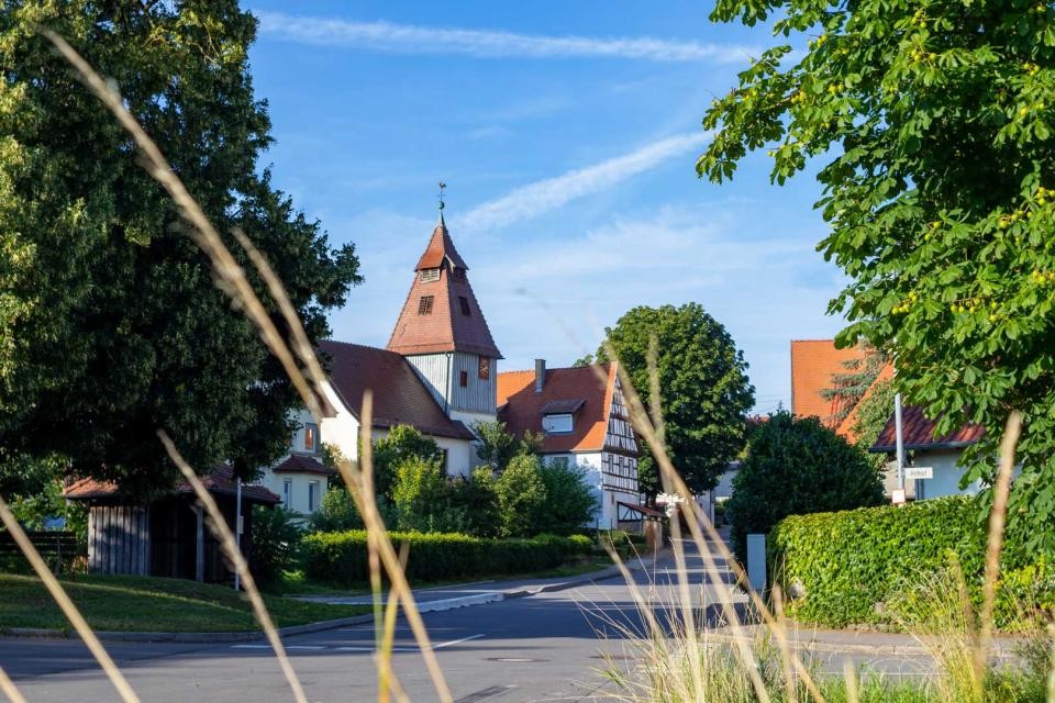Gemeinde Zweiflingen Rechts und Links stehen große grüne Laubbäume. In der Mitte führt eine Straße durch den Ort. Am linken Straßenrand steht eine kleine Dorfkirche und daneben ein Fachwerkhaus. Im Vordergrund ragen einzelne Grashalme in den Himmel.