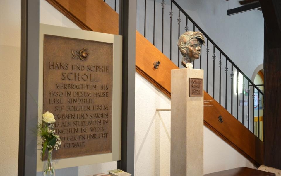 Erinnerung Sophie Scholl Gedenktafel von Hans und Sophie Scholl ist an zwei Säulen befestigt. Davor steht eine weiße Rose in einer Glasvase. Rechts daneben ist auf einer Steinsäule eine Büste des Kopfs von Sophie Scholl befestigt. Im Hintergrund führt eine Treppe aus Holz mit schwarzem Geländer nach oben.