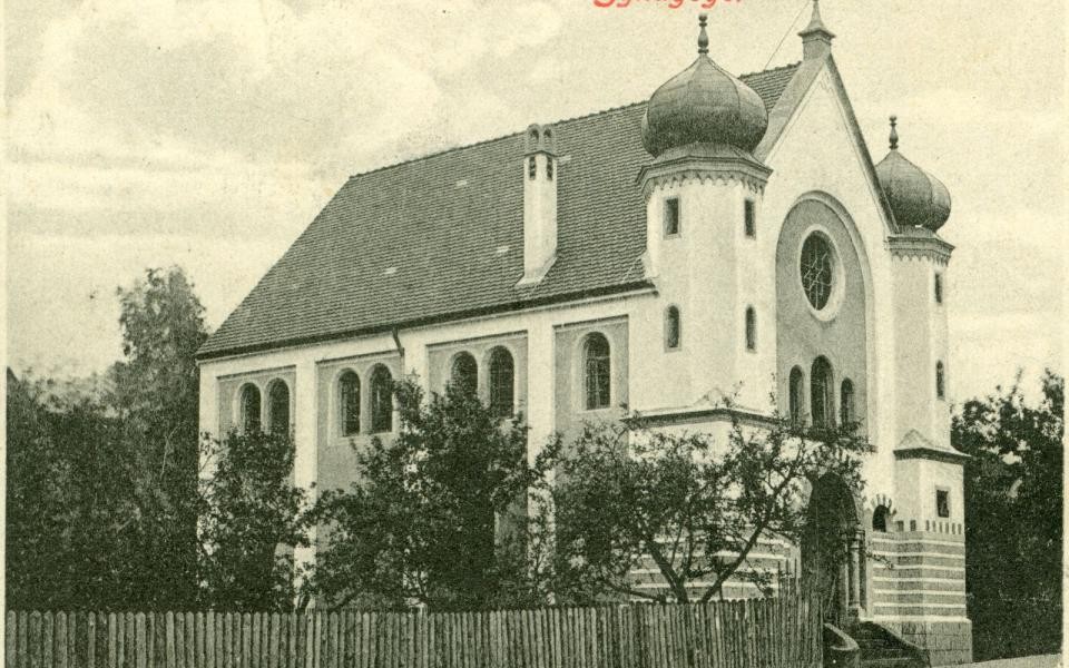 Die Synagoge ist ein Kirchengebäude neben der Eingangstüre befinden sich zwei Türme mit Kuppeldach. Der Eingang liegt direkt an einer Straße. Auf der linken Seite der Kirche geht an der Straße ein Holzzaun entlang. Dahinter befinden sich Bäume.