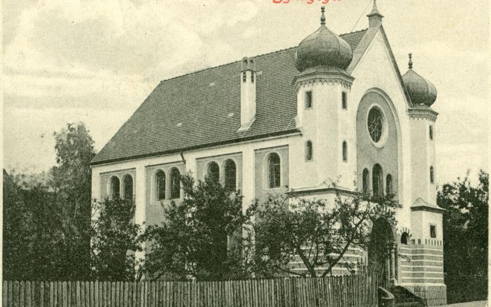 Synagoge Künzelsau Die Synagoge ist ein Kirchengebäude neben der Eingangstüre befinden sich zwei Türme mit Kuppeldach. Der Eingang liegt direkt an einer Straße. Auf der linken Seite der Kirche geht an der Straße ein Holzzaun entlang. Dahinter befinden sich Bäume.