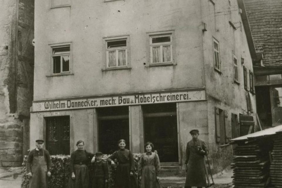 Möbelschreinerei in Neuenstein Ein schwarz-weiß Bild zeigt ein Wohnhaus im Erdgeschoss befindet sich eine Schreinerei. Vor dem Gebäude stehen Personen, die die Familie und das damalige Personal darstellen.