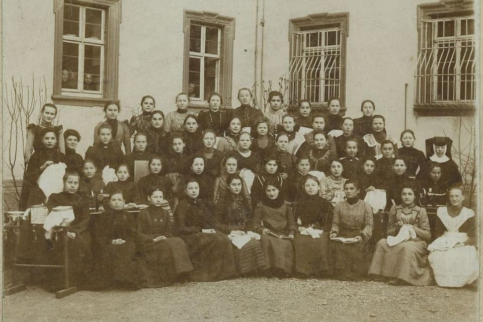 Gruppenbild Nähschule in Mulfingen ca. 50 Frauen in Kleider sitzen und stehen in 5 Reihen zusammen für ein Gruppenbild. Das Bild wurde vor einer Gebäudefassade aufgenommen. Im Hintergrund sind 4 Fenster.