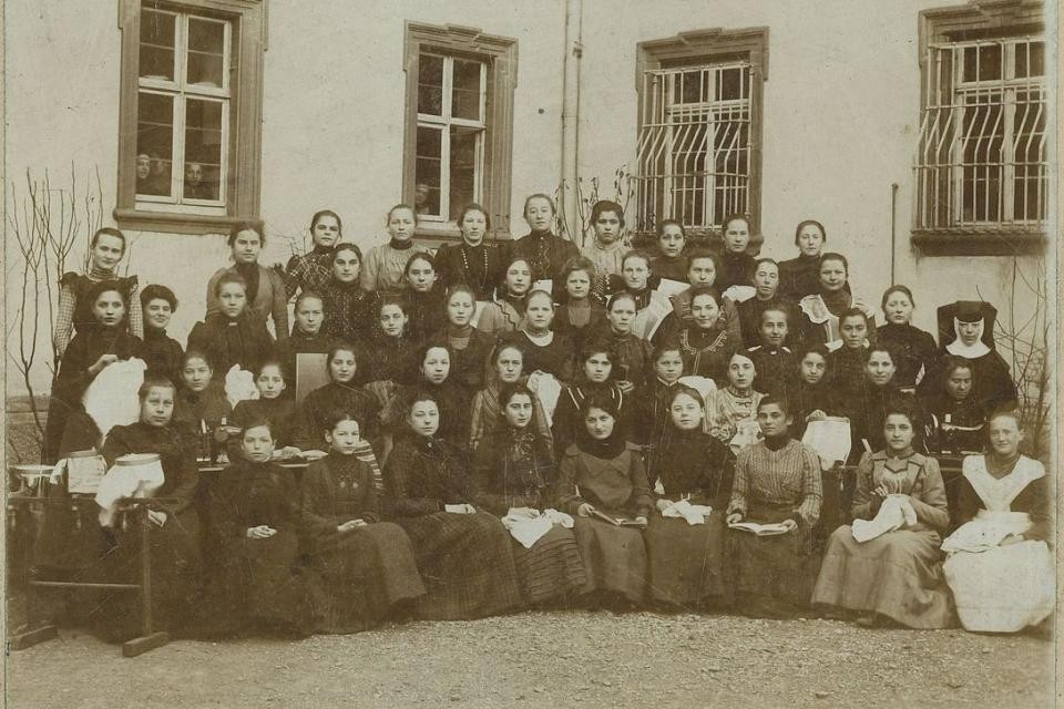 ca. 50 Frauen in Kleider sitzen und stehen in 5 Reihen zusammen für ein Gruppenbild. Das Bild wurde vor einer Gebäudefassade aufgenommen. Im Hintergrund sind 4 Fenster.