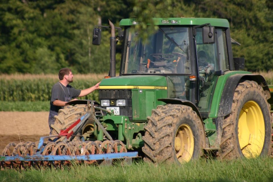 Ein grüner Traktor mit gelben Felgen steht auf einem Acker. Daneben steht ein junger Landwirt. Im Hintergrund steht auf einem Feld Mais, dahinter sind Bäume.