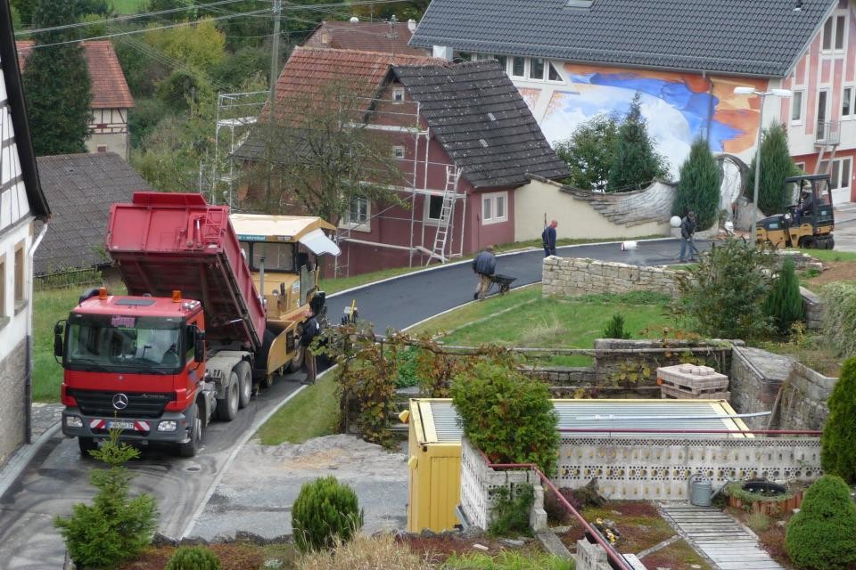Dorfflurneuordnung Eine Ortsdurchfahrt, die asphaltiert wird. Auf beiden Straßenseiten sind Grundstücke mit Häusern oder Gärten. Auf der Straße steht ein roter LKW; der gerade Material für die Asphaltierarbeiten abkippt.