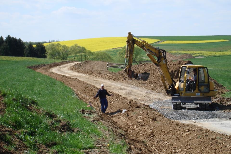 Feldwegebau Im Hintergrund befinden sich grüne und gelbe Felder und ein paar Bäume. Im Vordergrund steht ein gelber Bagger auf einen Feldweg, der aktuell gerichtet und ausgebaut wird. Ein Mitarbeiter mit Helm läuft im Graben.