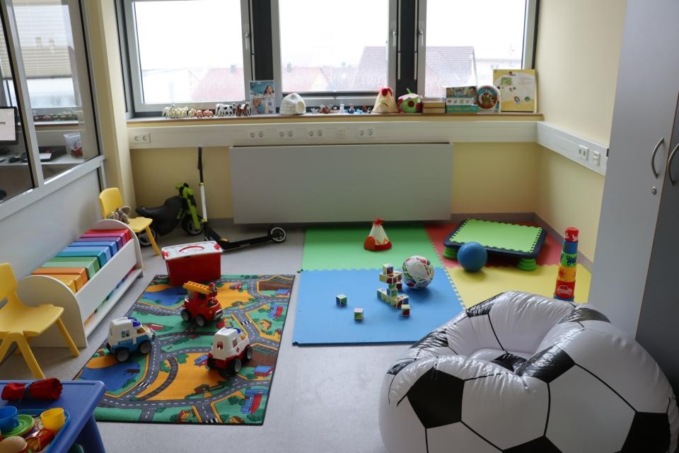 In einem Raum liegen Teppiche und Matten auf dem Boden und Spielsachen sind zu sehen. Auf der linken Seite sieht man ein Regal, ein kleiner gelber Stuhl und ein kleiner Tisch; auf der rechten Seite ist im vorderen Bildbereich ein Sitzsack zu sehen, der wie ein Fußball aussieht.