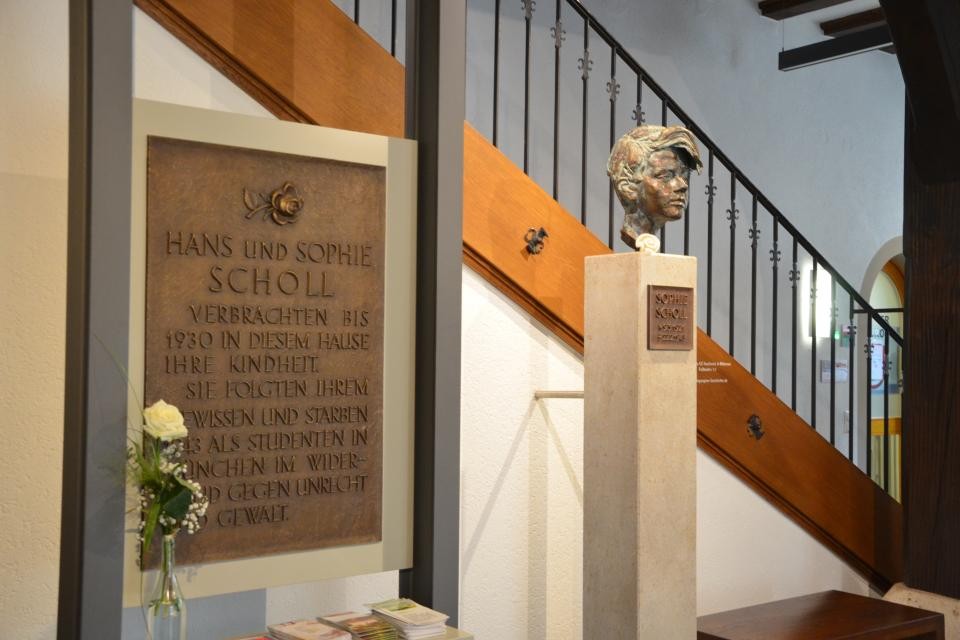 Gedenktafel von Hans und Sophie Scholl ist an zwei Säulen befestigt. Davor steht eine weiße Rose in einer Glasvase. Rechts daneben ist auf einer Steinsäule eine Büste des Kopfs von Sophie Scholl befestigt. Im Hintergrund führt eine Treppe aus Holz mit schwarzem Geländer nach oben.