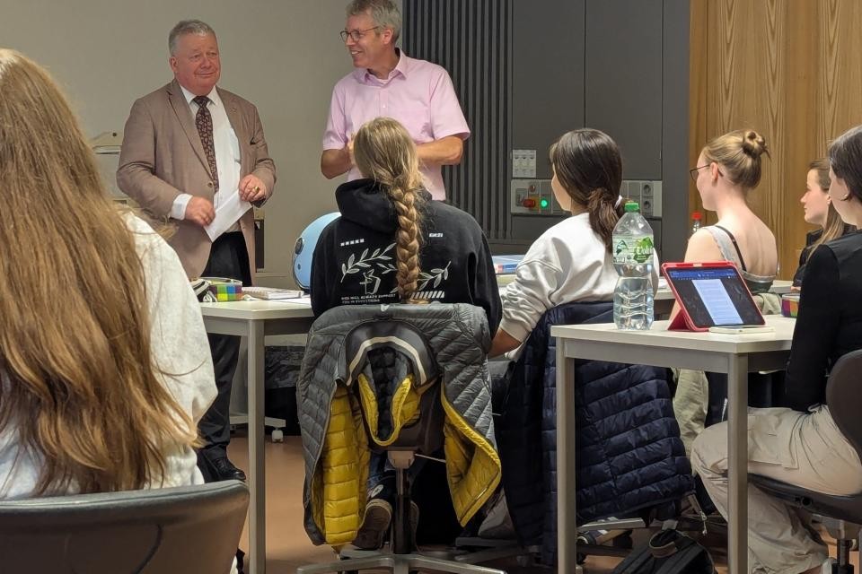 In der unteren Bildhälfte sind sitzende Schüler mit dem Rücken zur Kamera, mittig hinten stehen von Eyb und Maisborn zur Kamera gewandt.
