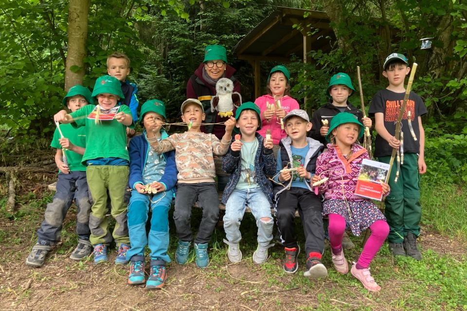 Elf Kinder in bunten Klamotten mit grünen Hüten im Vordergrund, sitzend als Gruppe. Im Hintergrund ist grüner Wald.