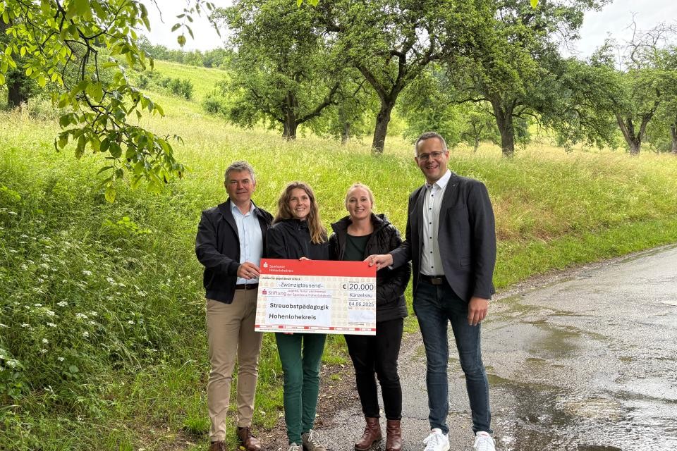 Im Vordergrund stehen links und rechts zwei Männer und in der Mitte zwei Frauen zur Kamera gewandt, die zwei Frauen mit einem rot-weißen Scheck in der Hand über 20.000 Euro. Im hintergrund sieht man grüne Bäume und die Streuobstwiese.
