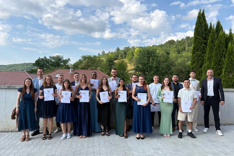Im Vordergrund auf der grau gepflasterten Terrasse sind Abiturienten und Abiturientinnen in festlicher Kleidung, alle halten ihre Zeugnisse hoch. Im Hintergrund blauer Himmel mit weißen Wolken und grüne Bäume.