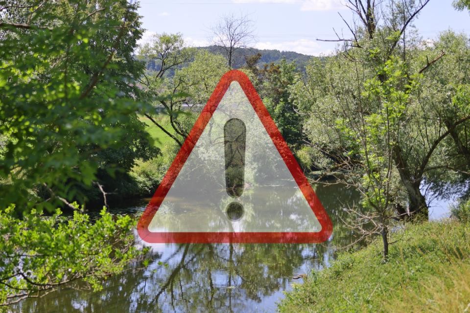 Im Vordergrund ist ein rot umrandetes Dreieck mit schwarz aufgemaltem Ausrufezeichen, leicht durchsichtig. Im Hintergrund sind grüne Wiesen und Bäume sowie ein Fluss.