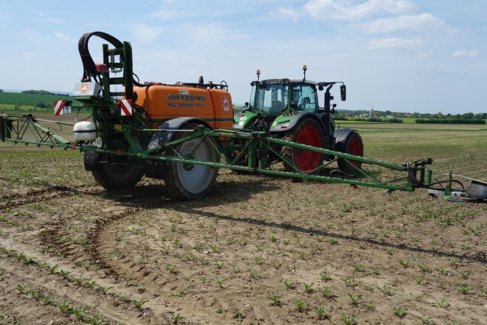 Braunes Feld, was von einem grünen Traktor mit orangefarbener Spritzmaschine bearbeitet wird.
