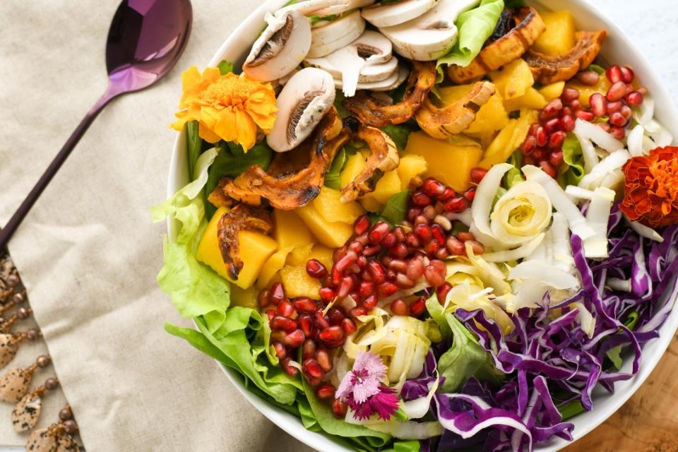 Weißer Teller mit buntem Obst und Gemüse drauf, wie Rotkohl, Salat, Mango, Champignons, Granatapfel und Zwiebeln. Links daneben ein lilafarbener Löffel. Darunter eine beige Tischdecke aus Leinen.