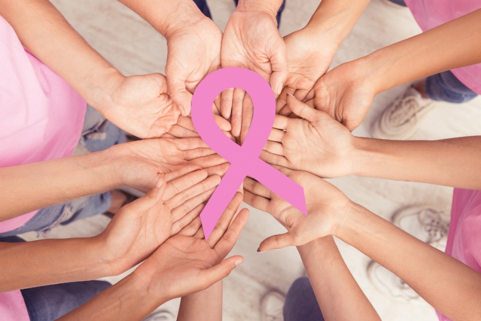 Viele Hände, die einen gemeinsamen Kreis formen. In der Mitte ist ein pinkes Zeichen für Krebshilfe. Die weiblichen Personen haben alle weiße Sneaker, blaue Jeans und ein rosa T-Shirt an. Perspektive von oben.