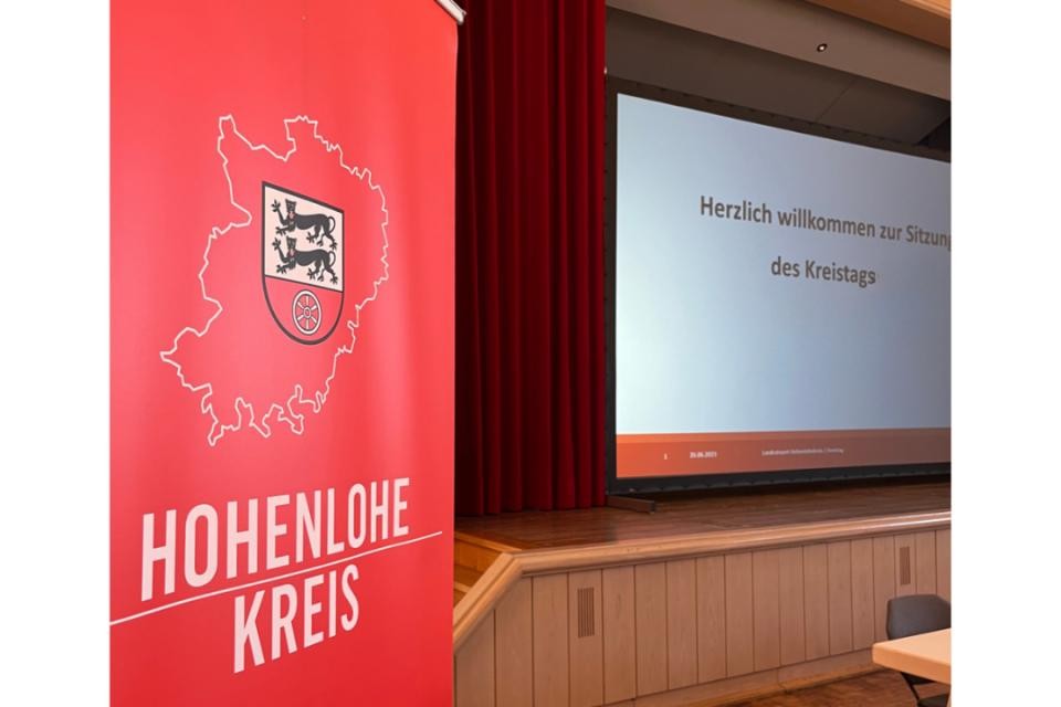 Roter Banner mit weißer Schrift "Hohenlohekreis". Dahinter ein weißer Bildschirm mit schwarzer Aufschrift "Herzlich Willkommen zur Sitzung des Kreistags"