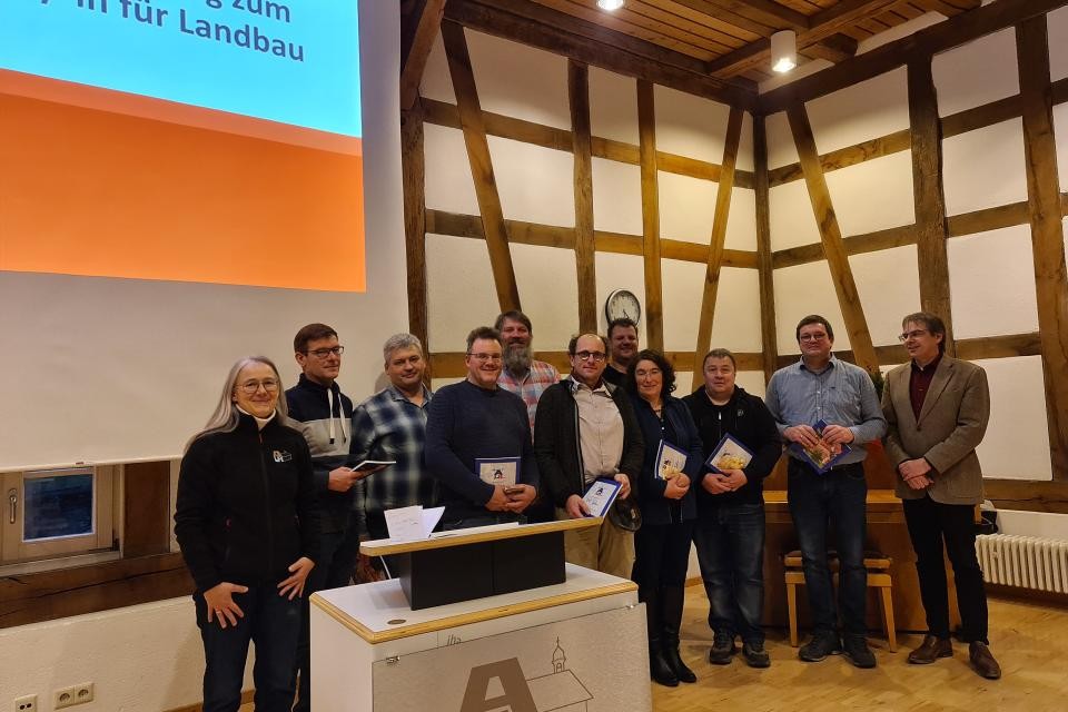 Eine Gruppe von Menschen steht hinter einem Podium. Sie halten weiße Urkunden in der Hand. Im Hintergrund ist eine weiße Wand mit Holz zu sehen.