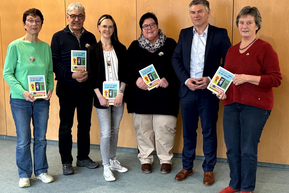 Eine Gruppe von Personen steht vor einer braunen Wand und hält bunte Flyer in den Händen.
