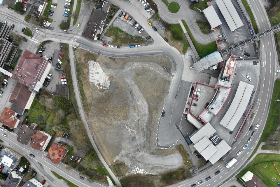 Eine große freie Fläche aus grauem Schotter. Links und rechts befibden sich asphaltierte Straßen und Häuser.