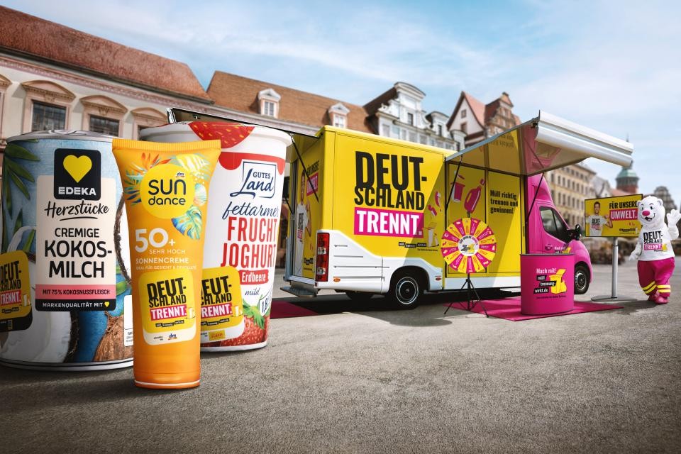 Große bunte XXl-Verpackungen der Produkte "Edeka Herzstücke cremige Kokosmilch", "Sun Dance 50+ Sonnencreme" und "Gutes Land Fruchtjoghurt" links auf grauem Asphalt aufgestellt. Rechts befindet sich der grell gelb-pinke Aktionswagen und Maskottchen "Trenn-Bär".