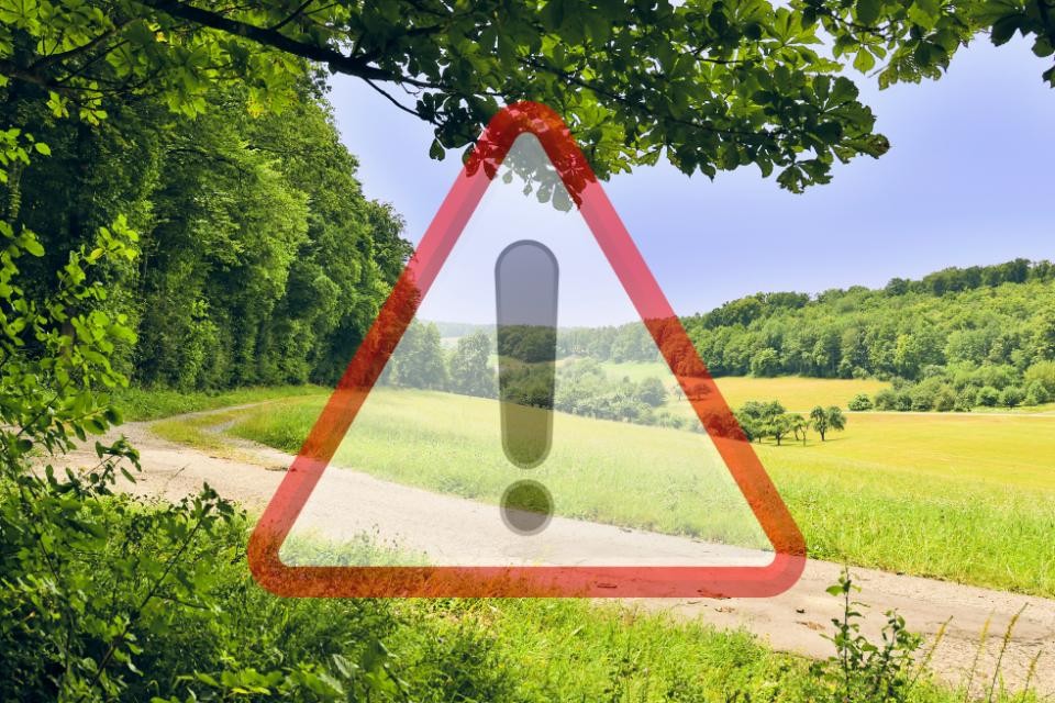 Ein geschotterter Feldweg führt durch eine grüne Wiese. Links ist ein grüner Wald. Im Hintergrund befindet sich blauer Himmel und ein Waldstück. Im Vordergrund befindet sich ein rot-weißes Warnschild mit einem Ausrufezeichen.
