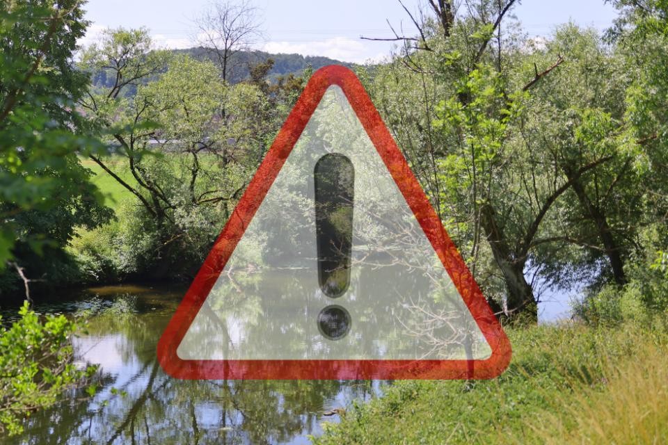 Ein Fluss umgeben von grünen Bäumen und Wiesen. Im Vordergrund ist ein weiß-rotes Warnsymbol mit schwarzem Ausrufezeichen. Im Hintergund ist Wald und blauer Himmel.