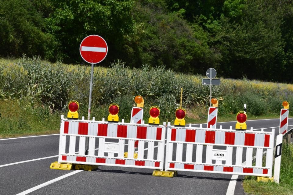 Sperrung einer Straße mit rot-weißen Absperrungen und einem Schild mit Durchfahrtsverbot. Im Hintergrund befindet sich ein günes Feld und ein Wald.