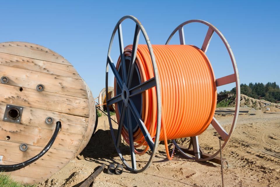 Zwei große Kabeltrommeln mit organgenem Kabel stehen auf einer Baustelle. Im Hintergrund ist blauer Himmel.