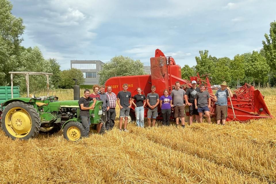 Eine Gruppe von Personen steht vor einem roten Mähdrescher und einem grünen Traktor auf einem gedroschenen Feld. Im Hintergrund sind grüne Bäume und blauer Himmell.