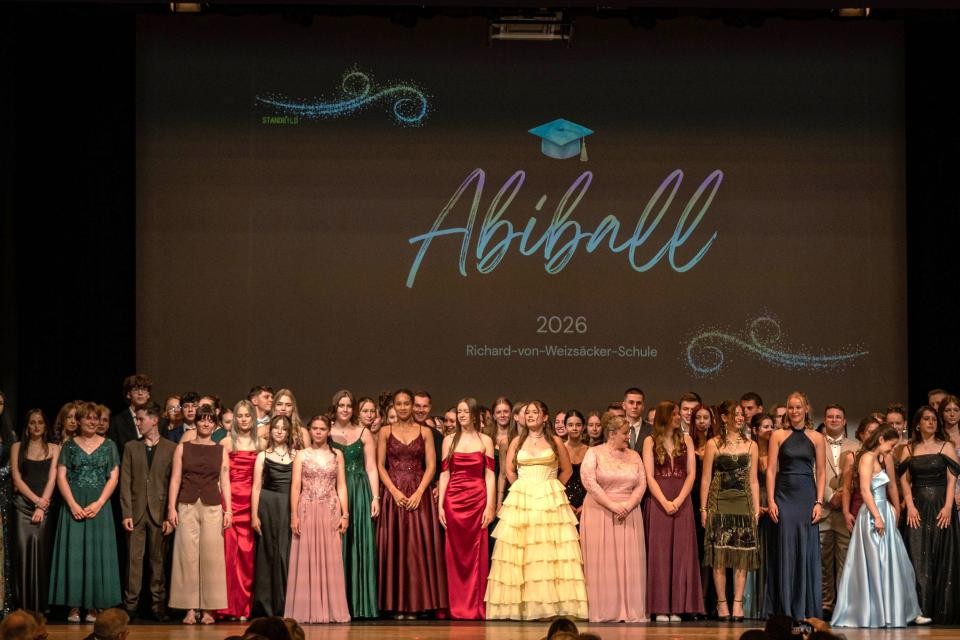 Eine Gruppe von Personen in bunten Kleidern steht auf einer Bühne. Im Hintergrund ist eine schwarze Leinwand mit der Aufschrift "Abiball 2025 Richard-von-Weizsäcker-Schule"