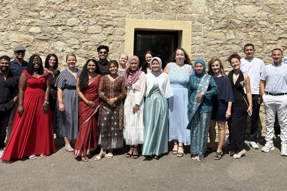 Eine Gruppe von Personen in festlicher Kleidung steht vor einer Gebäudemauer aus grau-braunem Stein.