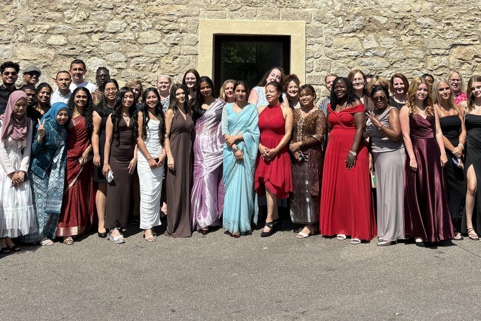 Eine Gruppe von Personen in festlicher Kleidung steht vor einer Gebäudemauer aus grau-braunem Stein.