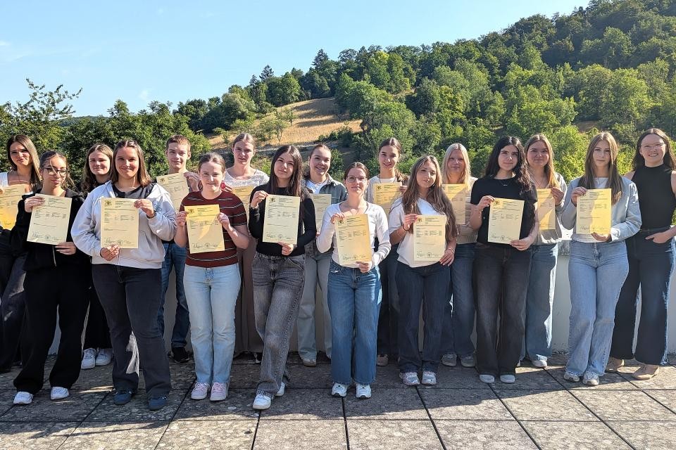 Eine Gruppe von Personen hält ihre Zeugnisse in der Hand. Im Hintergrund befindet sich ein grüner Wald und blauer Himmel.